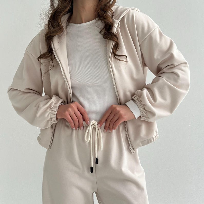 May include: A beige two-piece tracksuit featuring a zip-up hooded jacket and matching drawstring pants. The jacket has a white undershirt visible underneath. The outfit is made of a soft, comfortable material.