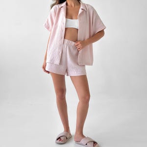May include: A light pink, short-sleeved button-down shirt and matching shorts set. The fabric has a textured, crinkled appearance. The model is also wearing a white top and light pink slippers.