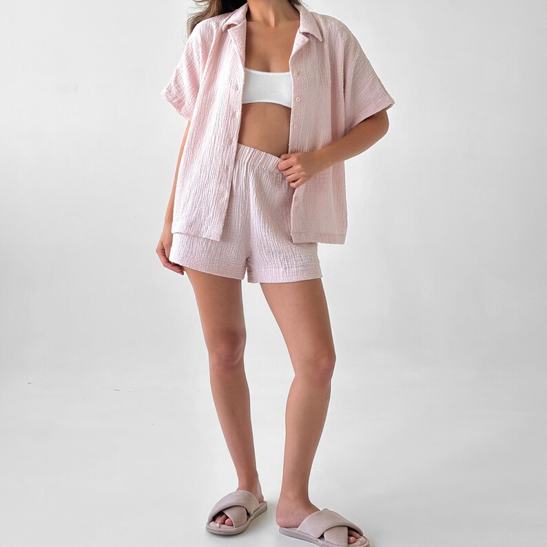 May include: A light pink, short-sleeved button-down shirt and matching shorts set. The fabric has a textured, crinkled appearance. The model is also wearing a white top and light pink slippers.