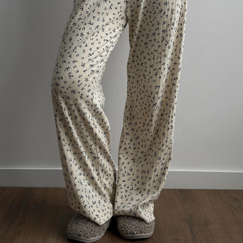 May include: Cream-colored, wide-leg pants with a small, dark floral pattern. The pants are paired with gray, fuzzy slippers. The fabric appears to be ribbed. The outfit is casual and comfortable.