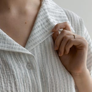 May include: Close-up of a light blue and white striped button-down shirt. The shirt has a textured, crinkled fabric and a classic collar. A hand is touching the collar, and a small ring is visible on the finger.