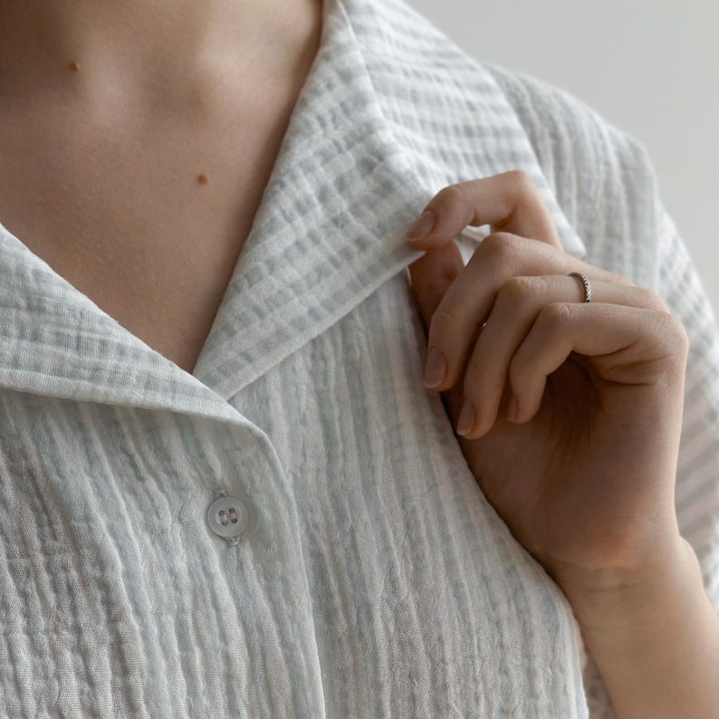 May include: Close-up of a light blue and white striped button-down shirt. The shirt has a textured, crinkled fabric and a classic collar. A hand is touching the collar, and a small ring is visible on the finger.