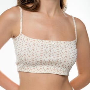 Op de afbeelding: Cr&egrave;mekleurige crop top met een bloemenpatroon in roze en bruine tinten. De top heeft dunne bandjes en een klein strikje in het midden. De stof lijkt geribbeld.
