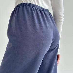 May include: Close-up of blue sweatpants with an elastic waistband. The pants are a solid color and appear to be made of a soft material. A white long-sleeve shirt is partially visible in the background.