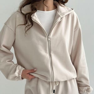 May include: A beige zip-up hoodie with a matching drawstring bottom. The hoodie has a hood and ribbed cuffs. The outfit is made of a soft material. The zipper is silver.