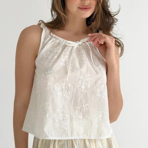 May include: A sleeveless white top featuring a delicate eyelet embroidery pattern with floral motifs and wavy lines. The garment has a gathered neckline with a thin white drawstring tie at the center, creating a soft, draped silhouette.