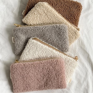 May include: A collection of six rectangular, fluffy pouches in various neutral colors: brown, beige, gray, white, and pink. Each pouch has a gold-colored zipper closure. The pouches are made of a soft, textured material.
