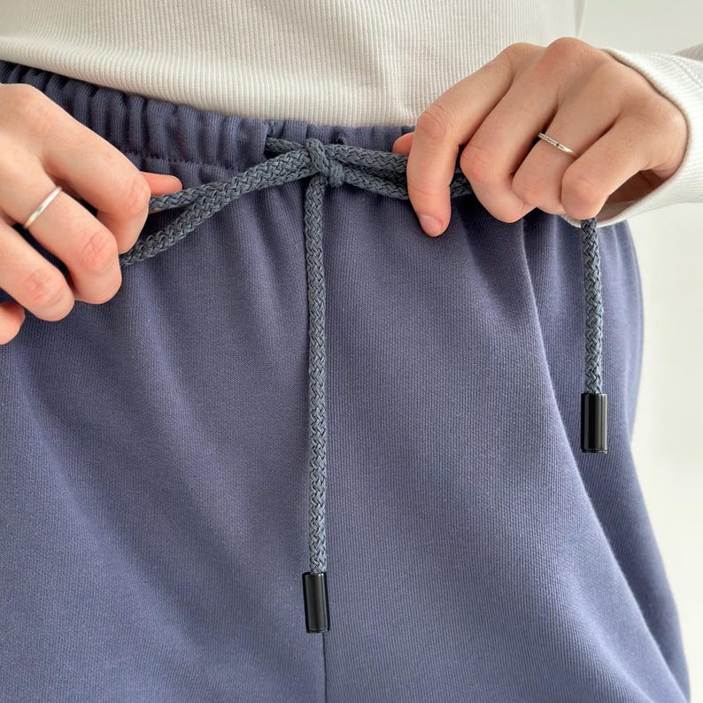 May include: Close-up of blue sweatpants with a gray drawstring. The drawstring has black aglets. The pants are being held by a person wearing a white long-sleeve shirt and a silver ring.