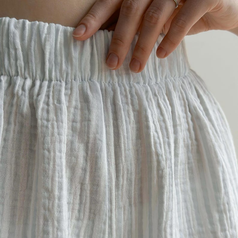 May include: Close-up of white, textured fabric, possibly a skirt or pants, with an elastic waistband. The fabric has vertical lines and a slightly crinkled appearance. A hand with a ring is resting on the waistband.