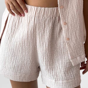 May include: Light pink shorts and a matching button-up shirt made of a textured fabric. The shorts have an elastic waistband. The shirt has three buttons. The fabric has a subtle, dotted pattern.