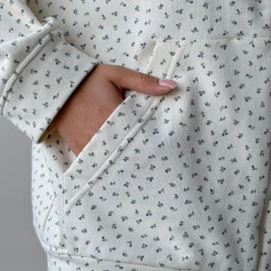 Op de afbeelding: Cr&egrave;mekleurige hoodie met een bloemenpatroon. De stof heeft kleine blauwe en groene bloemen verspreid over het oppervlak. De hoodie heeft een voorzak en een capuchon met trekkoord, wat een casual en comfortabele stijl biedt.