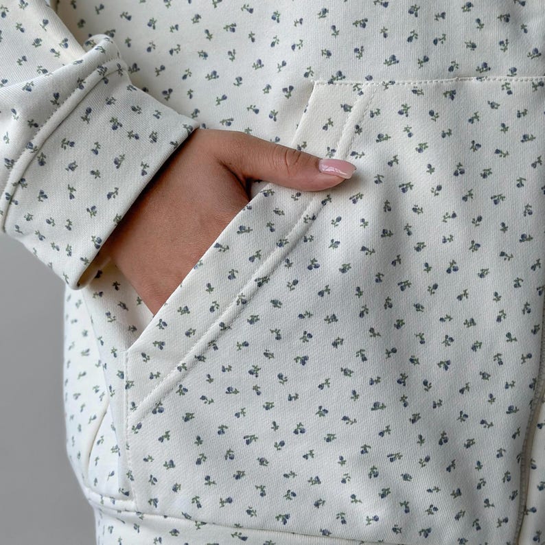 Op de afbeelding: Cr&egrave;mekleurige hoodie met een bloemenpatroon. De stof heeft kleine blauwe en groene bloemen verspreid over het oppervlak. De hoodie heeft een voorzak en een capuchon met trekkoord, wat een casual en comfortabele stijl biedt.
