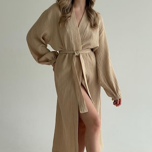 May include: A beige, long-sleeved robe with a V-neck and a matching belt tied at the waist. The robe has a textured appearance and a high slit on the left side, creating a flowing silhouette. The sleeves are long and loose.