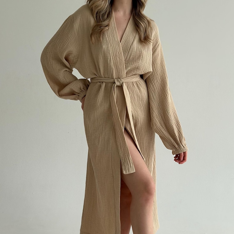 May include: A beige, long-sleeved robe with a V-neck and a matching belt tied at the waist. The robe has a textured appearance and a high slit on the left side, creating a flowing silhouette. The sleeves are long and loose.