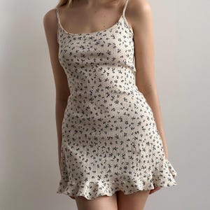 May include: A cream-colored, ribbed knit dress with a floral pattern. The dress features thin spaghetti straps and a ruffled hem. The small, dark blue floral print covers the entire dress, creating a delicate and feminine look. The dress is a mini length.