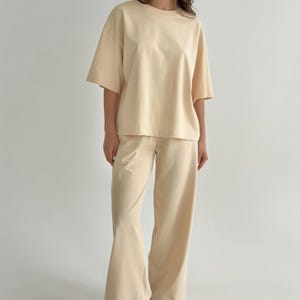 Könnte beinhalten: Ein passendes zweiteiliges Set in einfarbigem Hellbeige. Das Set besteht aus einem kurzärmeligen T-Shirt mit Rundhalsausschnitt und lockerer, kastiger Passform sowie einer langen Hose mit weitem Bein und geradem Schnittverlauf.