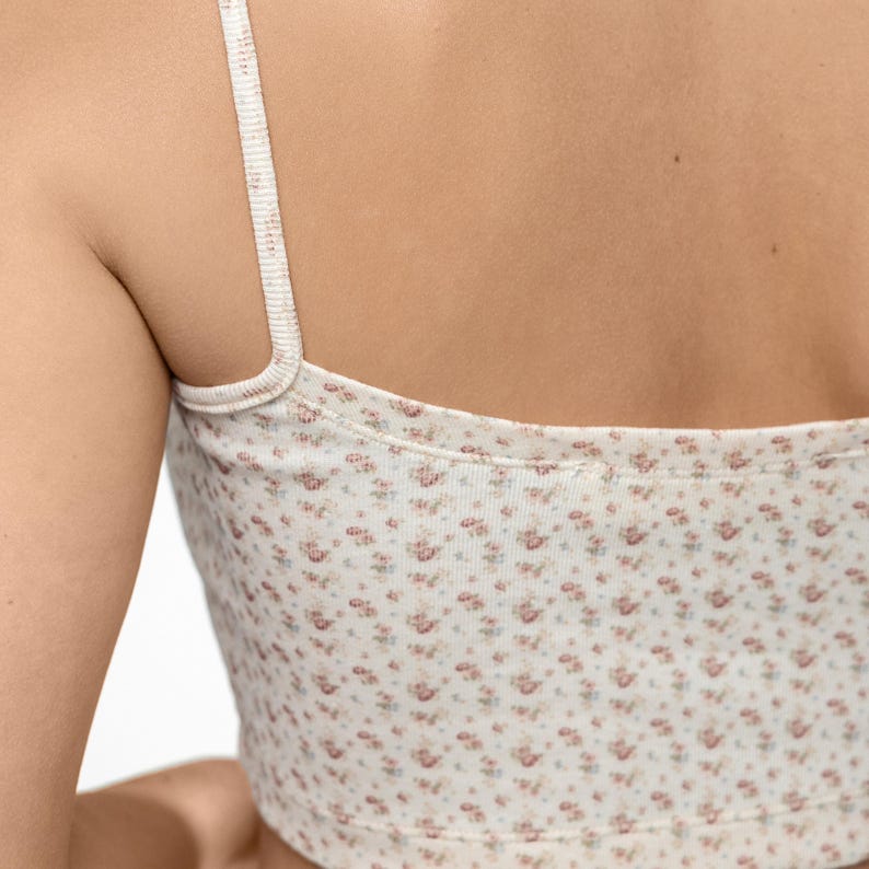 Op de afbeelding: Een cr&egrave;mekleurige crop top met een delicaat bloemenpatroon in roze en rode tinten. De top heeft dunne bandjes en een ronde halslijn. De stof lijkt zacht en licht, geschikt voor warm weer.