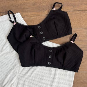 May include: Two black ribbed bralettes featuring thin adjustable straps with small pink rose accents, a scoop neckline, and a front placket with three black buttons. The bralettes are layered over a pair of white ribbed shorts with a subtle textured pattern.