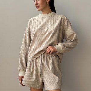 Puede incluir: Conjunto de sal&oacute;n beige de dos piezas con una parte superior de manga larga y pantalones cortos. La parte superior tiene cuello redondo y pu&ntilde;os acanalados, mientras que los pantalones cortos tienen cintura con cord&oacute;n. La tela tiene un patr&oacute;n texturizado y punteado.
