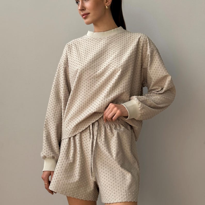Puede incluir: Conjunto de sal&oacute;n beige de dos piezas con una parte superior de manga larga y pantalones cortos. La parte superior tiene cuello redondo y pu&ntilde;os acanalados, mientras que los pantalones cortos tienen cintura con cord&oacute;n. La tela tiene un patr&oacute;n texturizado y punteado.