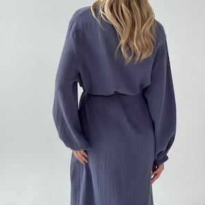 May include: A long-sleeved, blue-grey dress with a cinched waist and a flowing skirt. The dress has a textured fabric and a relaxed fit. The sleeves are long and gathered at the wrists. The model is wearing the dress from the back.