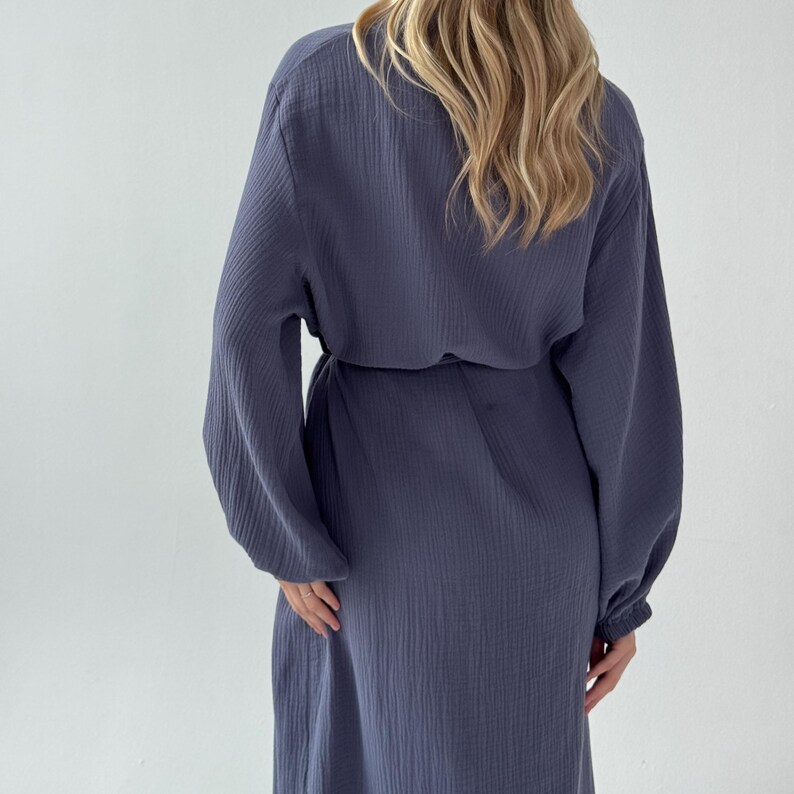 May include: A long-sleeved, blue-grey dress with a cinched waist and a flowing skirt. The dress has a textured fabric and a relaxed fit. The sleeves are long and gathered at the wrists. The model is wearing the dress from the back.