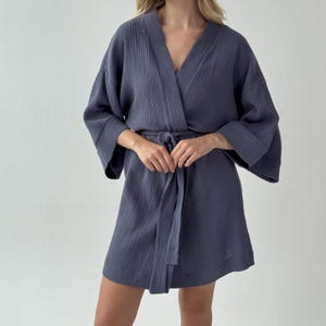 May include: A short, slate blue robe with a V-neck and wide sleeves. The robe is tied at the waist with a matching fabric belt. The fabric has a textured, crinkled appearance. The robe is a comfortable garment.