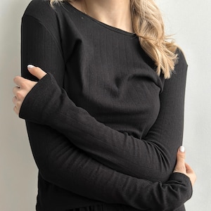 May include: Black long-sleeved top with a ribbed texture. The top features a scoop neckline and fitted sleeves. The fabric looks soft and comfortable. The sleeves are long and cover the arms.