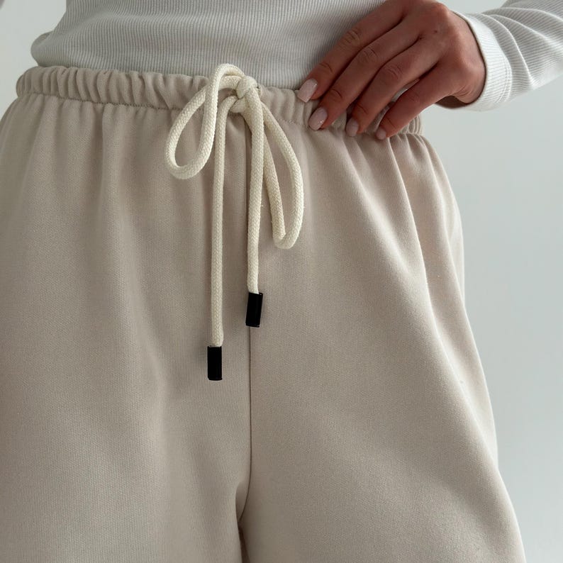 May include: Close-up of beige sweatpants with a drawstring. The drawstring is white with black aglets. The waistband is elastic. The pants are made of a soft, comfortable material.
