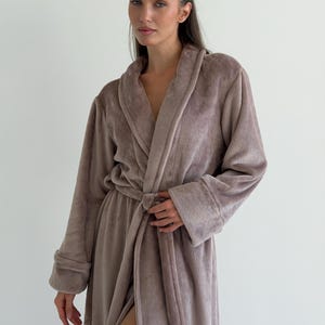 Könnte beinhalten: Ein flauschiger Bademantel in Taupe mit Schalkragen, langen Ärmeln mit umgeschlagenen Bündchen und einem passenden Bindegürtel aus Stoff in der Taille. Das Kleidungsstück besteht aus einem weichen, flauschigen Material mit gleichmäßiger Textur.