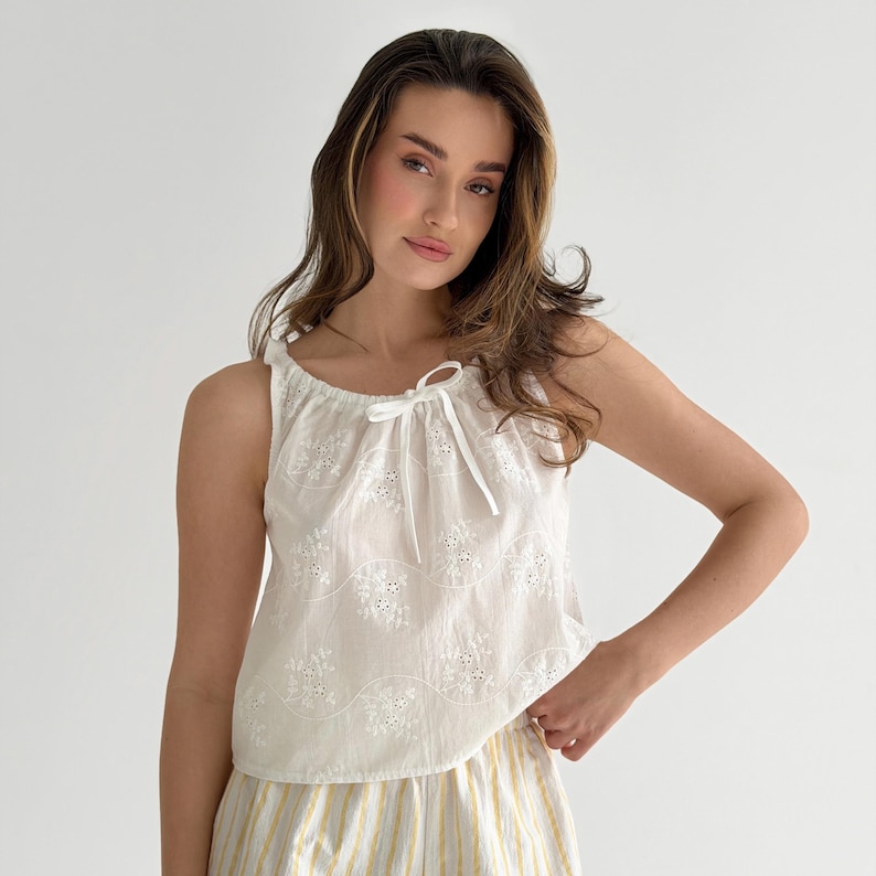 May include: A sleeveless white top featuring an eyelet lace floral pattern and a drawstring neckline with a tie closure. The garment is paired with white and yellow vertical striped bottoms, creating a coordinated two-piece set ensemble.