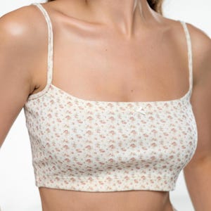 Op de afbeelding: Cr&egrave;mekleurige crop top met een bloemenpatroon in roze en groene tinten. De top heeft dunne bandjes en een vierkante halslijn. Een klein strikje is in het midden aan de voorkant geplaatst. De stof lijkt geribbeld.