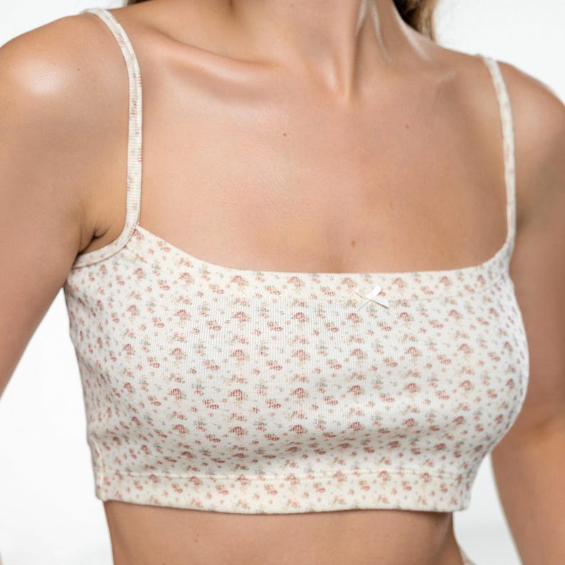 Op de afbeelding: Cr&egrave;mekleurige crop top met een bloemenpatroon in roze en groene tinten. De top heeft dunne bandjes en een vierkante halslijn. Een klein strikje is in het midden aan de voorkant geplaatst. De stof lijkt geribbeld.