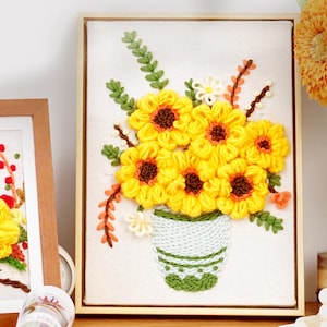 May include: Embroidered artwork featuring a bouquet of yellow sunflowers in a white vase with green accents. The flowers have brown centers and are surrounded by green leaves and stems. The artwork is framed in a light brown wooden frame.