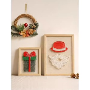 Puede incluir: Arte mural con temática navideña que presenta una corona, un regalo enmarcado y un diseño de Papá Noel enmarcado. Los diseños del regalo y de Papá Noel están hechos con arte de hilo. La corona está hecha de ramas tejidas, vegetación y adornos.