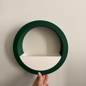 Puede incluir: Un estante de pared circular con un anillo exterior verde oscuro y un estante interior blanco. El estante está montado en una pared blanca. El estante está sostenido por una mano.