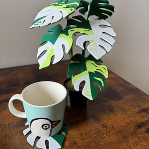 Könnte beinhalten: Eine grün-weiße Monstera-Pflanze mit einem Untersetzer mit weißem Vogelmotiv und einer hellblauen Tasse mit weißem Vogelmotiv.