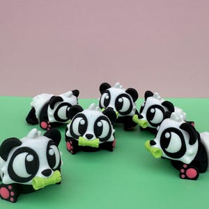 Puede incluir: Seis figuras de panda, con cuerpo blanco, marcas negras y pies rosas. Sostienen un brote de bambú verde. Los pandas están sobre una superficie verde, con un fondo rosa. Las figuras son pequeñas y detalladas.