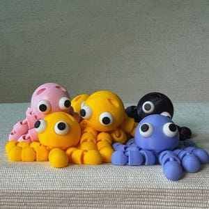 Puede incluir: Un grupo de juguetes de pulpo articulados y coloridos en rosa, amarillo, azul y negro. Cada pulpo tiene ojos grandes y saltones y tentáculos segmentados. Los juguetes están dispuestos sobre una superficie texturizada blanquecina.