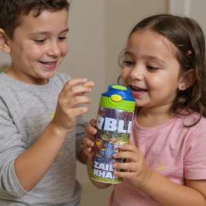 K&ouml;nnte beinhalten: Eine bunte Wasserflasche mit blauem, gr&uuml;nem und gelbem Deckel. Die Flasche hat ein lebendiges Grafikdesign mit dem Namen "Zail Khan" und dem Wort "BLI". Die Flasche wird von zwei Kindern gehalten.