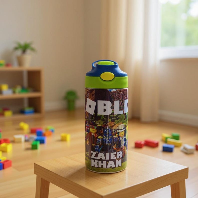 K&ouml;nnte beinhalten: Eine Wasserflasche mit blauem und gr&uuml;nem Deckel und einer farbenfrohen H&uuml;lle mit dem Wort "BL" und dem Namen "ZAIER KHAN". Die zylindrische Flasche steht auf einem kleinen Holztisch in einem Raum mit Spielzeug.