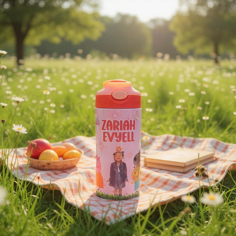 K&ouml;nnte beinhalten: Eine rosa-orangefarbene Wasserflasche mit dem Aufdruck "Zariah Evyell". Die Flasche zeigt eine Cartoon-Illustration eines M&auml;dchens und anderer Charaktere. Die Flasche steht auf einer Picknickdecke auf einer Wiese.