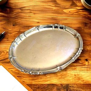 Può includere: Un vassoio ovale argentato con bordi smerlati ornati. Il vassoio è realizzato in metallo lucido e riflettente e si trova su una superficie di legno. Il design del vassoio è classico ed elegante, adatto per servire cibo o esporre oggetti.