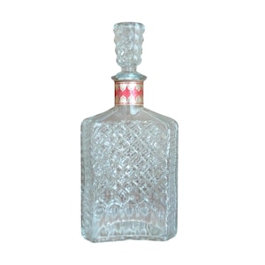 May include: A clear, square-shaped glass decanter with a diamond-patterned design. The decanter has a decorative stopper and a red and gold label around the neck. The bottle is empty and sits against a white background.