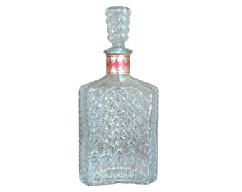 May include: A clear, square-shaped glass decanter with a diamond-patterned design. The decanter has a decorative stopper and a red and gold label around the neck. The bottle is empty and sits against a white background.