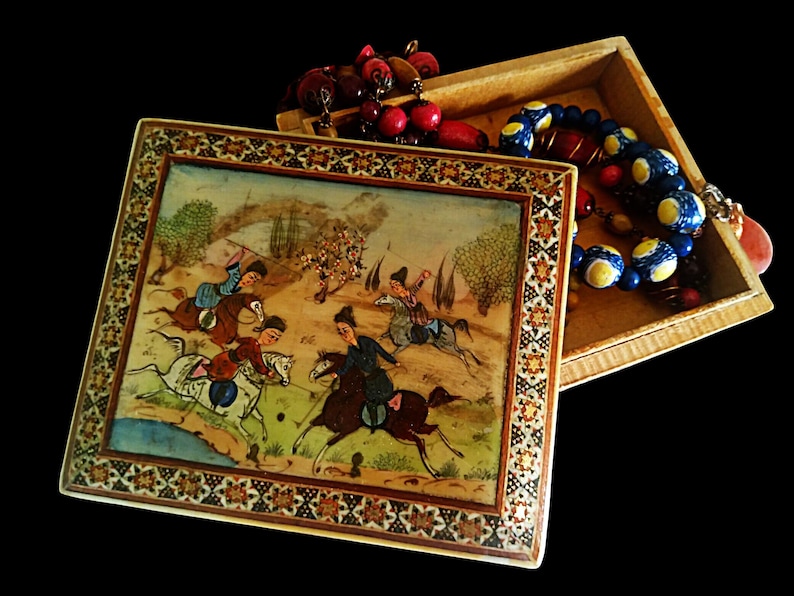 May include: An open wooden box with a hand-painted lid depicting a polo match scene. The box is decorated with an ornate border. Inside the box are several beaded necklaces in red, blue, and yellow. The background is black.
