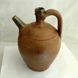 Può includere: Una brocca in ceramica marrone con beccuccio e manico. La brocca ha un corpo arrotondato e un beccuccio e collo verde scuro. Il manico è arcuato e attaccato alla parte superiore della brocca. La brocca sembra fatta a mano e ha un aspetto rustico.