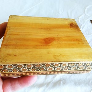 May include: A small, rectangular wooden box with a light brown finish. The box features a decorative border with a repeating geometric pattern in shades of brown and tan. The box is held in a hand.
