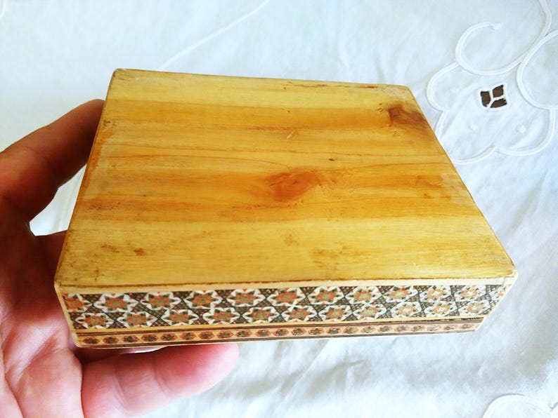 May include: A small, rectangular wooden box with a light brown finish. The box features a decorative border with a repeating geometric pattern in shades of brown and tan. The box is held in a hand.