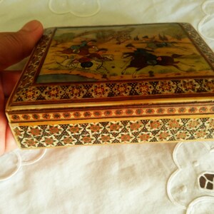 May include: A rectangular wooden box with a detailed painting on the lid depicting figures on horseback in a landscape. The box has intricate geometric patterns in shades of brown, gold, and cream. The box is held by a hand.